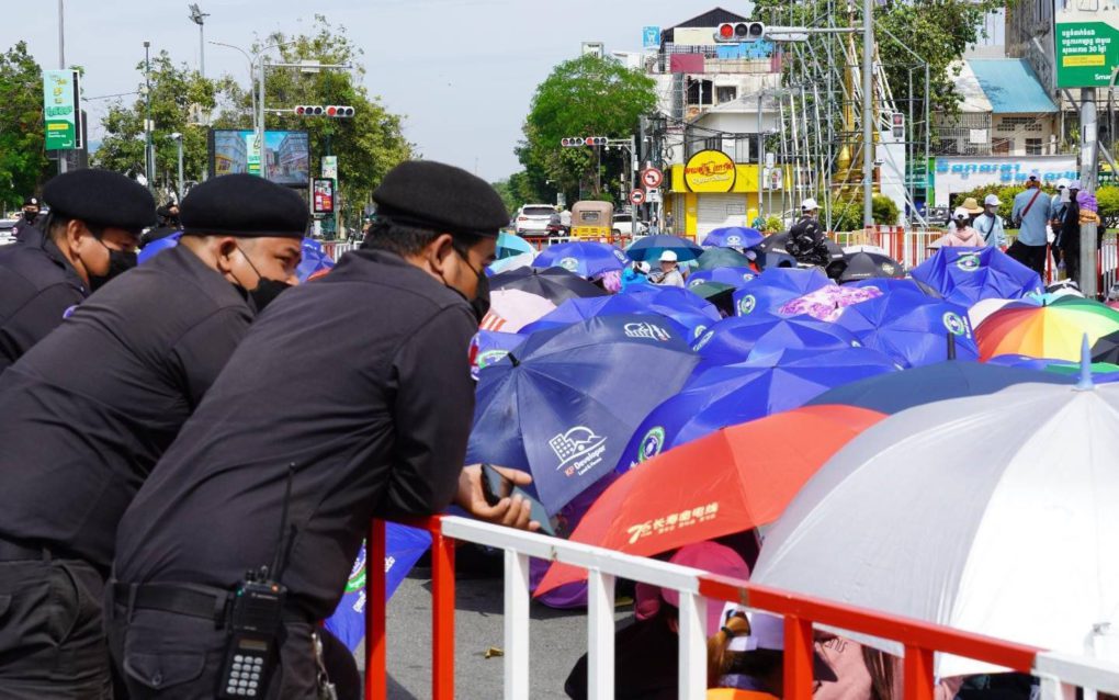 Police lean on a barricade and watch protesting workers take cover under umbrellas during the NagaWorld strike on December 19, 2021. (Tran Techseng/VOD)