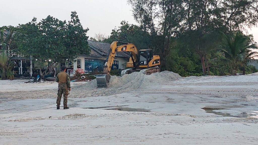 A uniformed guard watches an excavator dig into sand on Koh Rong Sanloem island's Saracen Bay resort area on February 4, 2023. (Danielle Keeton-Olsen/VOD)